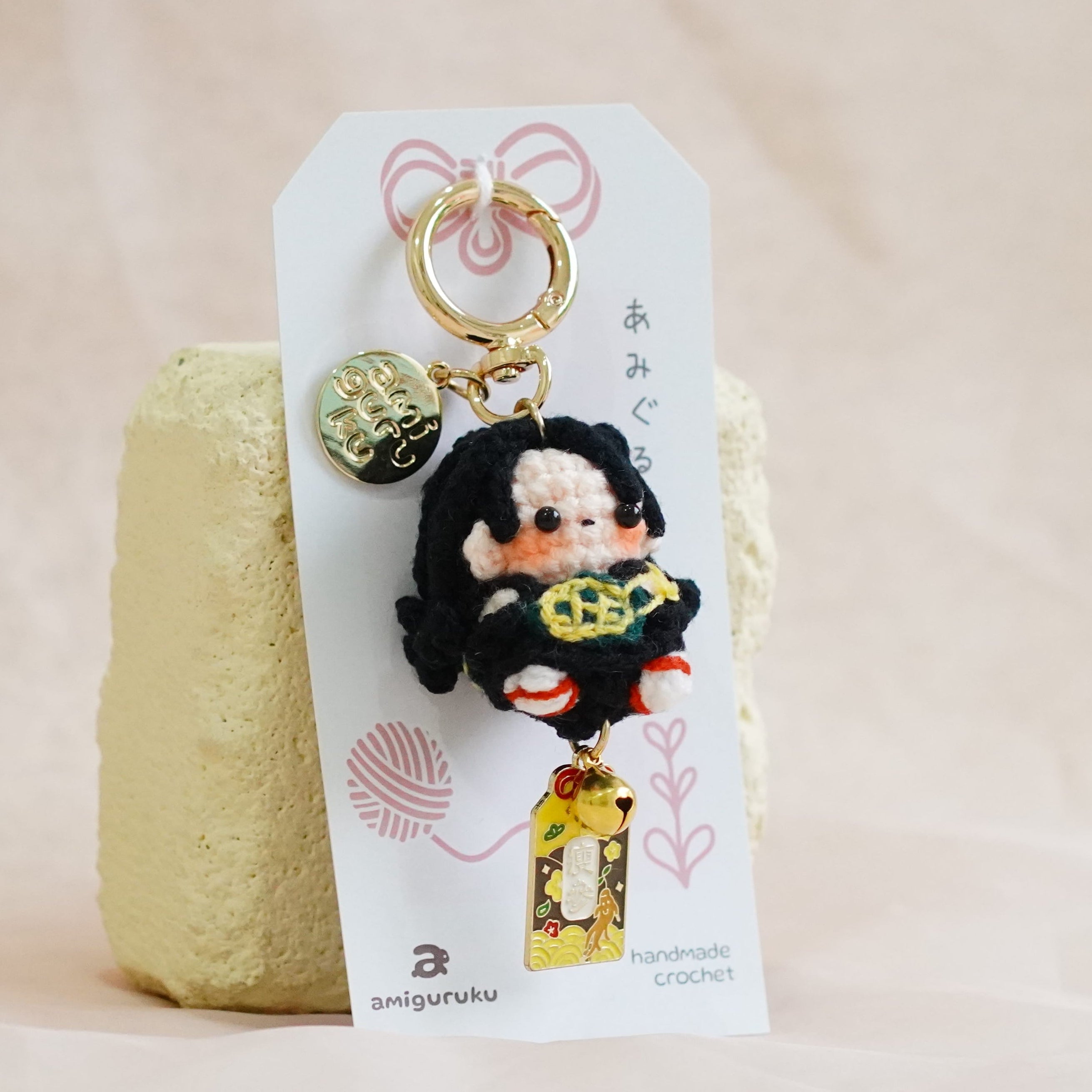 A small plush keychain or bag charm displayed on a white hanging card, resting against a textured pale yellow block on a soft beige fabric surface. The charm features a black-haired character in a black outfit holding a yellow lantern, with a gold key ring and small decorative charms.