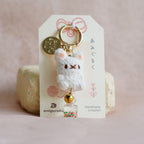 A handmade keychain featuring a small white crocheted bear with embroidered facial features, a gold key ring, a gold coin charm, and a small bell, displayed on a branded white card.