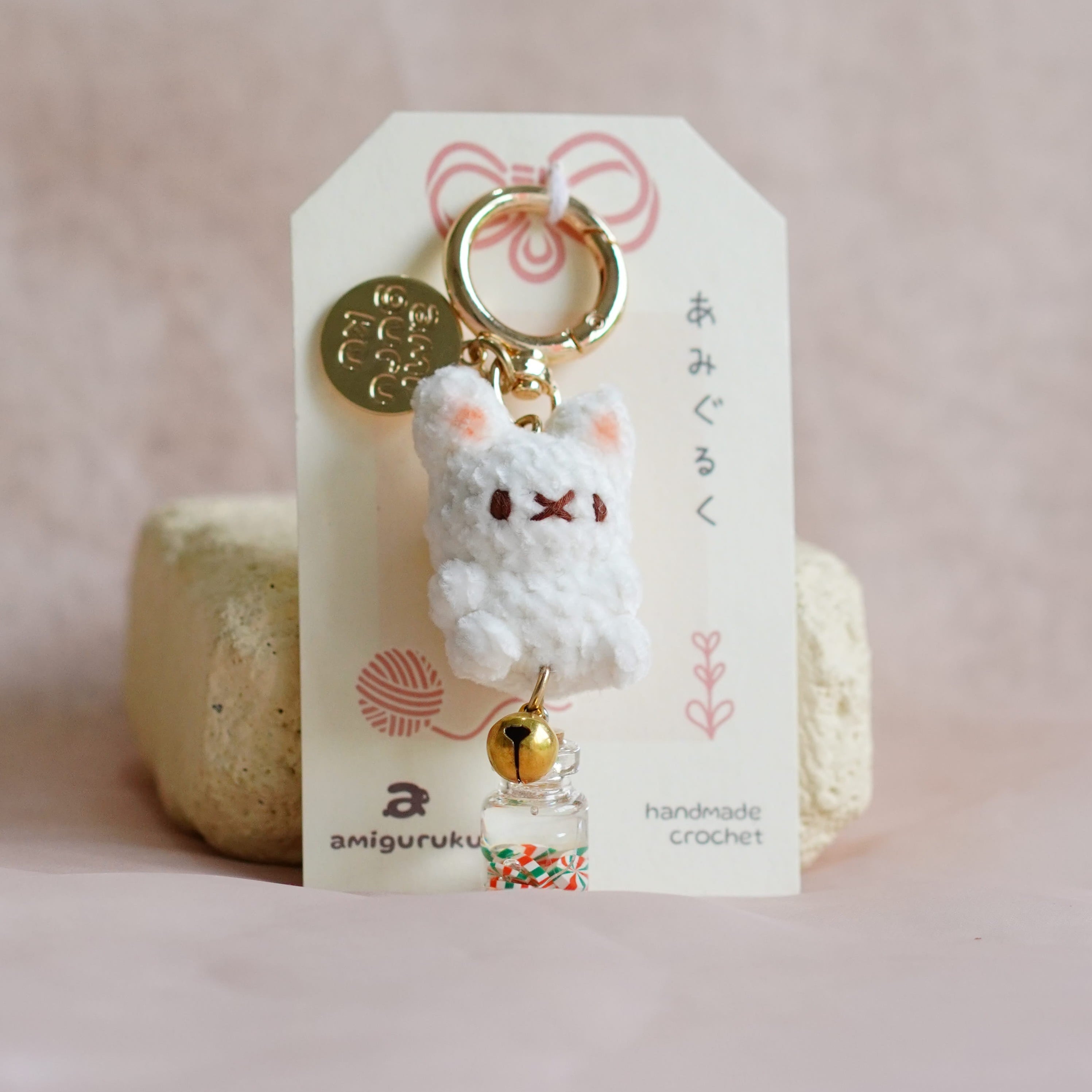 A handmade keychain featuring a small white crocheted bear with embroidered facial features, a gold key ring, a gold coin charm, and a small bell, displayed on a branded white card.