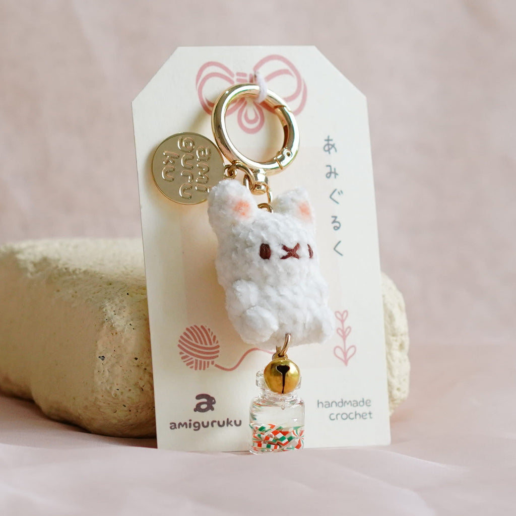 A small white plush charm keychain with gold-toned hardware and a printed display card, photographed on soft blush fabric with a beige cushion behind it.