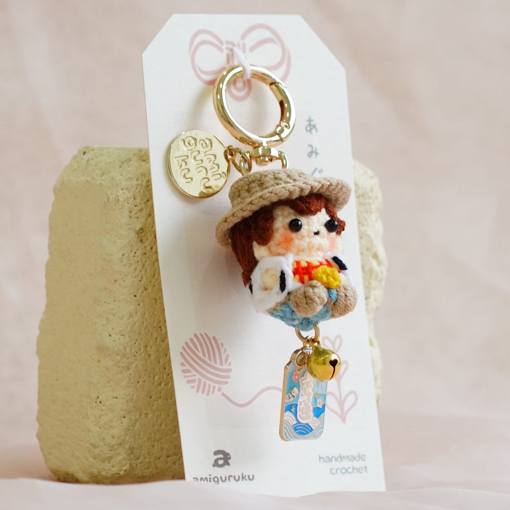 A small embroidered keychain charm displayed on a white hanging card, standing against a soft beige fabric backdrop with a textured beige block behind it.