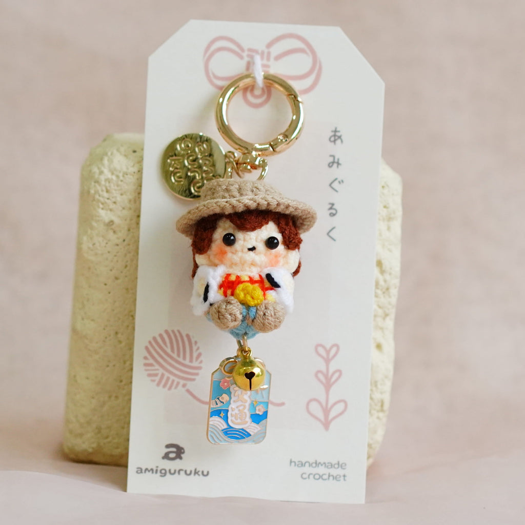 A handmade Japanese doll keychain charm displayed on its white paper packaging card, set against a soft beige fabric background.