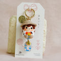 A handmade Japanese doll keychain charm displayed on its white paper packaging card, set against a soft beige fabric background.
