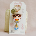 A handmade Japanese doll keychain charm displayed on its white paper packaging card, set against a soft beige fabric background.